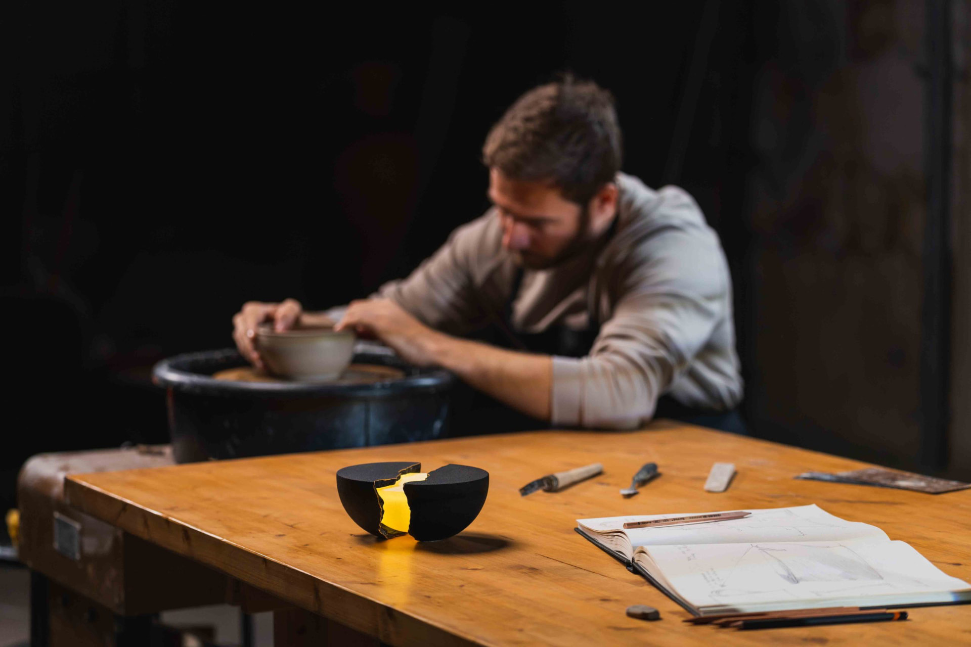 Glowing split-hemisphere lamp on a workbench while a potter works at a wheel in the background