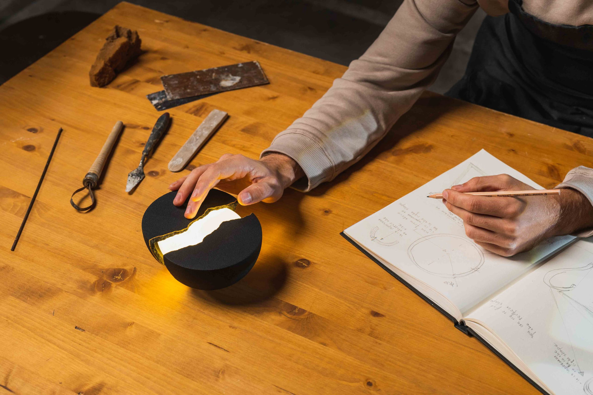 Overhead view of a designer's hands on a glowing split-hemisphere lamp beside an open sketchbook