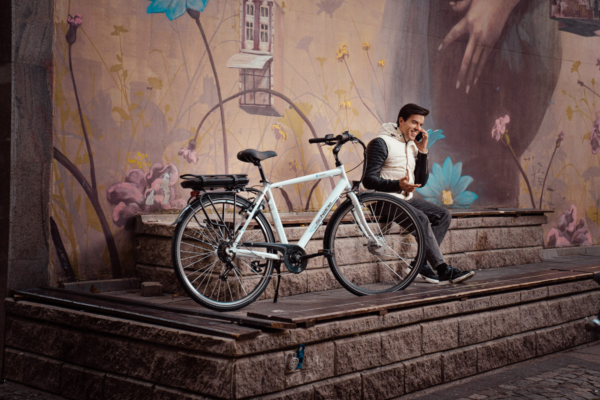 White electric city bike parked on stone steps beside a man talking on a phone in front of a mural