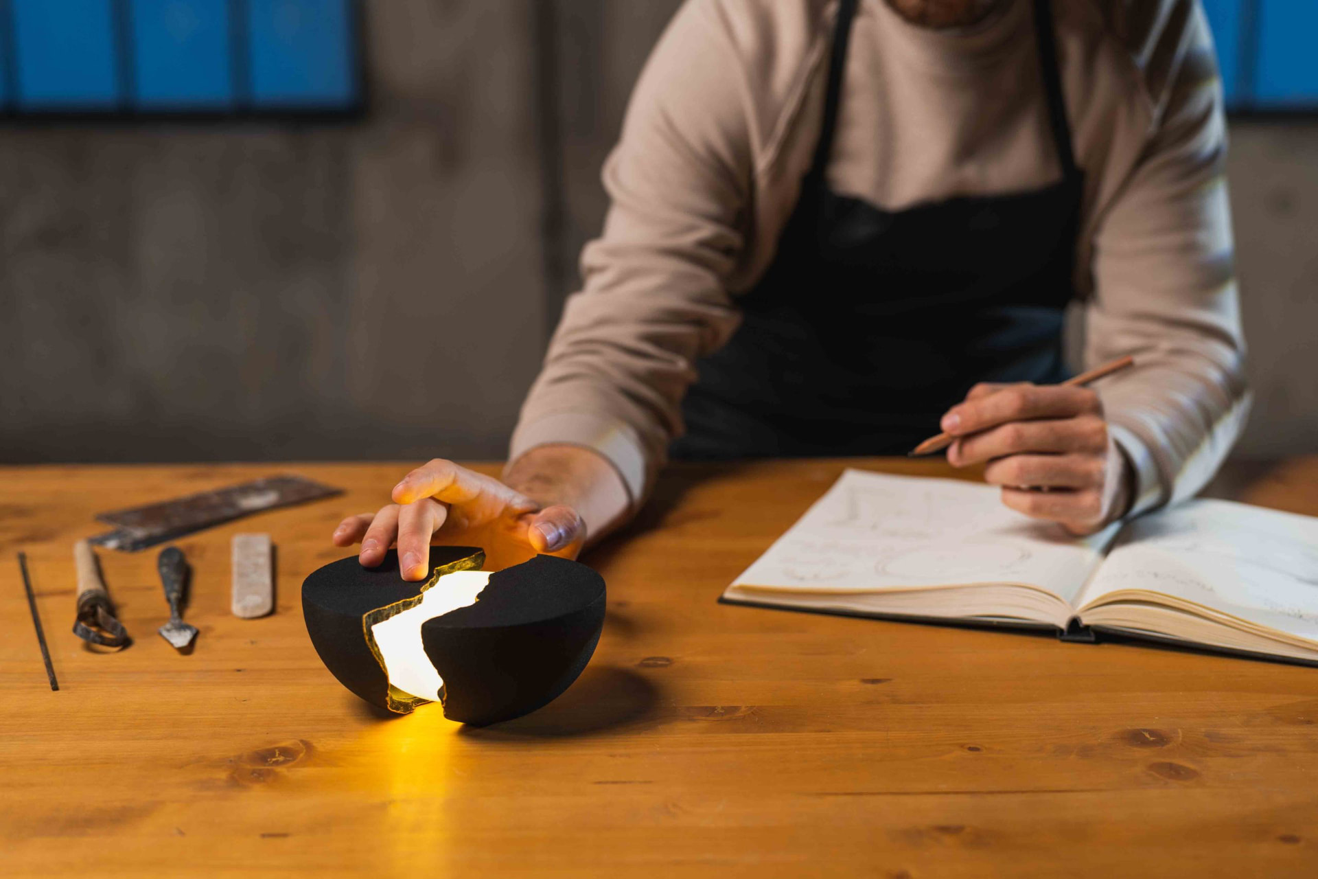 Craftsman sketching at a wooden workbench beside a split-hemisphere black lamp glowing amber