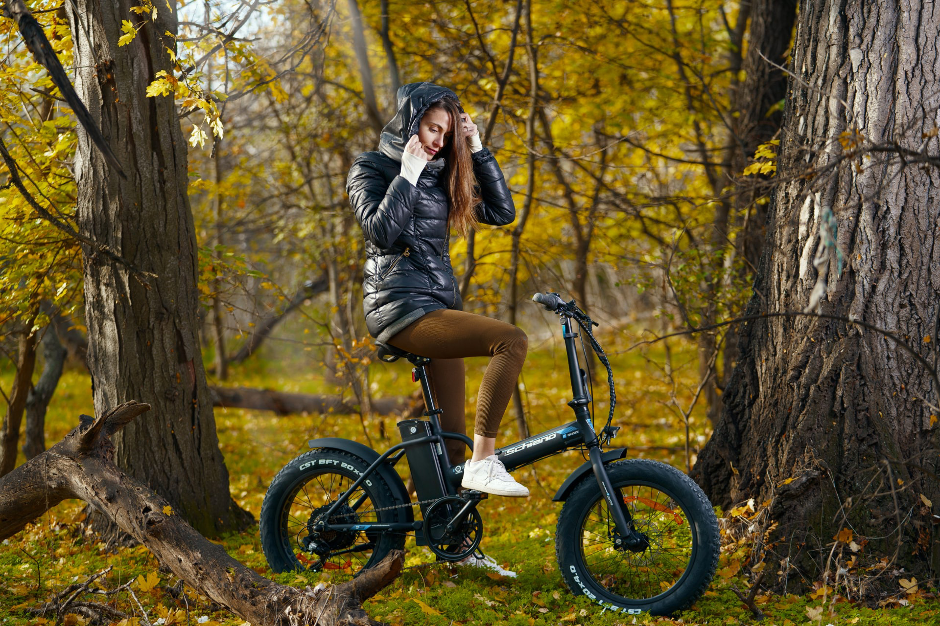 Woman straddling a black folding electric fat-tyre bike on an autumn forest floor